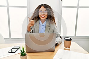 Beautiful hispanic business woman sitting on desk at office working with laptop doing happy thumbs up gesture with hand