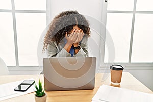 Beautiful hispanic business woman sitting on desk at office working with laptop with sad expression covering face with hands while