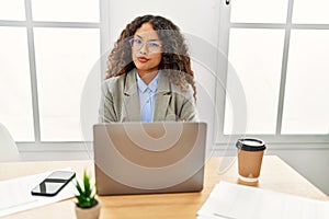 Beautiful hispanic business woman sitting on desk at office working with laptop skeptic and nervous, disapproving expression on