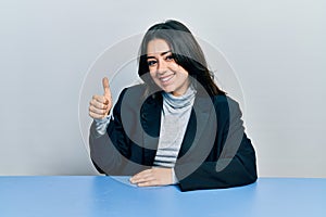 Beautiful hispanic business woman sitting on the table smiling happy and positive, thumb up doing excellent and approval sign