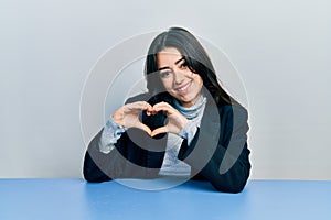 Beautiful hispanic business woman sitting on the table smiling in love showing heart symbol and shape with hands