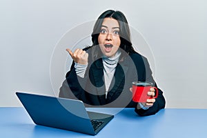 Beautiful hispanic business woman working at the office drinking a cup of coffee pointing thumb up to the side smiling happy with