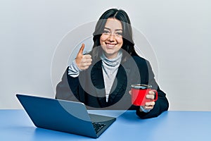 Beautiful hispanic business woman working at the office drinking a cup of coffee smiling happy and positive, thumb up doing