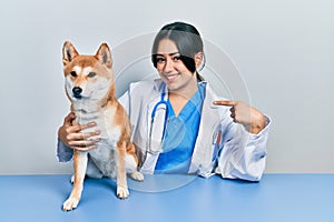 Beautiful hispanic veterinarian woman checking dog health pointing finger to one self smiling happy and proud