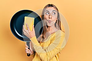 Beautiful hispanic woman cleaning cooking pan with scourer making fish face with mouth and squinting eyes, crazy and comical