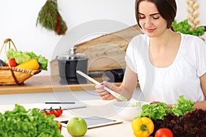 Beautiful Hispanic woman cooking in kitchen while using tablet computer and wooden spoon. Housewife found new recipe for