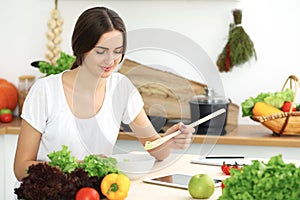 Beautiful Hispanic woman cooking in kitchen while using tablet computer and wooden spoon. Housewife found new recipe for