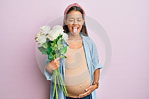 Beautiful hispanic woman expecting a baby holding flowers sticking tongue out happy with funny expression