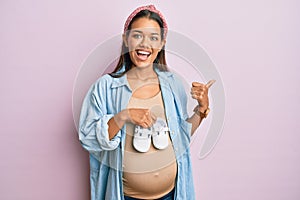 Beautiful hispanic woman expecting a baby holding shoes pointing thumb up to the side smiling happy with open mouth