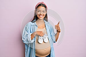 Beautiful hispanic woman expecting a baby holding shoes smiling happy and positive, thumb up doing excellent and approval sign