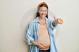 Beautiful hispanic woman expecting a baby, touching pregnant belly approving doing positive gesture with hand, thumbs up smiling