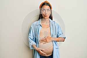 Beautiful hispanic woman expecting a baby, touching pregnant belly making fish face with lips, crazy and comical gesture