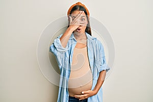 Beautiful hispanic woman expecting a baby, touching pregnant belly peeking in shock covering face and eyes with hand, looking
