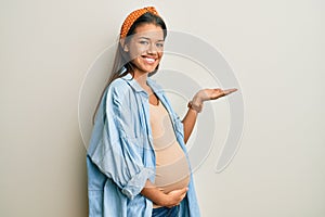 Beautiful hispanic woman expecting a baby, touching pregnant belly pointing aside with hands open palms showing copy space,