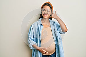 Beautiful hispanic woman expecting a baby, touching pregnant belly smiling doing phone gesture with hand and fingers like talking