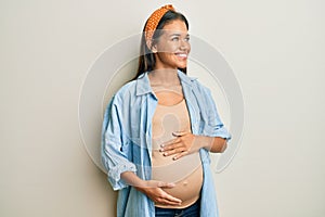 Beautiful hispanic woman expecting a baby, touching pregnant belly smiling looking to the side and staring away thinking