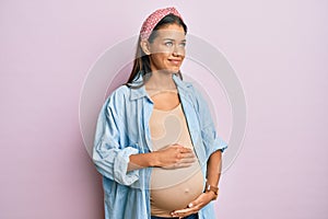 Beautiful hispanic woman expecting a baby, touching pregnant belly smiling looking to the side and staring away thinking