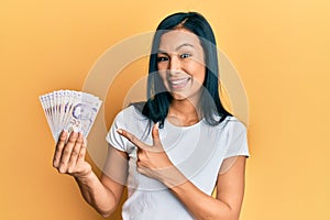 Beautiful hispanic woman holding 2 singapore dollars banknotes smiling happy pointing with hand and finger