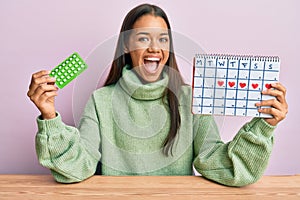 Beautiful hispanic woman holding birth control pills celebrating crazy and amazed for success with open eyes screaming excited