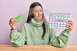 Beautiful hispanic woman holding birth control pills clueless and confused expression