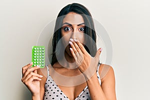 Beautiful hispanic woman holding birth control pills covering mouth with hand, shocked and afraid for mistake