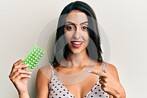 Beautiful hispanic woman holding birth control pills smiling happy pointing with hand and finger