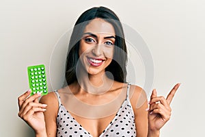 Beautiful hispanic woman holding birth control pills smiling happy pointing with hand and finger to the side