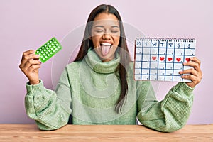 Beautiful hispanic woman holding birth control pills sticking tongue out happy with funny expression