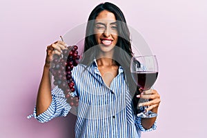 Beautiful hispanic woman holding branch of fresh grapes and red wine sticking tongue out happy with funny expression