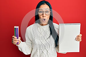 Beautiful hispanic woman holding canvas book and colored pencils afraid and shocked with surprise and amazed expression, fear and