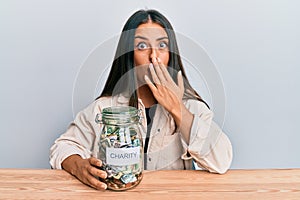 Beautiful hispanic woman holding charity jar with money covering mouth with hand, shocked and afraid for mistake