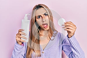 Beautiful hispanic woman holding cotton pad and make up remover in shock face, looking skeptical and sarcastic, surprised with