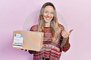 Beautiful hispanic woman holding delivery package pointing thumb up to the side smiling happy with open mouth