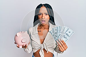 Beautiful hispanic woman holding dollars and piggy bank afraid and shocked with surprise and amazed expression, fear and excited
