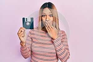 Beautiful hispanic woman holding floppy disk covering mouth with hand, shocked and afraid for mistake