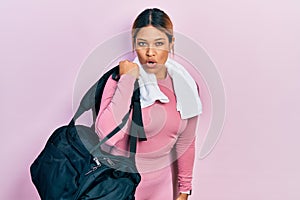 Beautiful hispanic woman holding gym bag and towel scared and amazed with open mouth for surprise, disbelief face