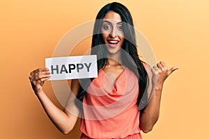 Beautiful hispanic woman holding happy text pointing thumb up to the side smiling happy with open mouth