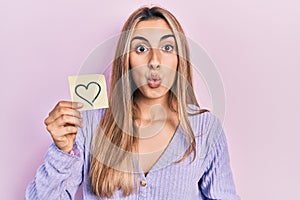 Beautiful hispanic woman holding heart reminder scared and amazed with open mouth for surprise, disbelief face