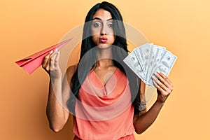 Beautiful hispanic woman holding paper plane and dollars puffing cheeks with funny face