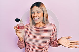 Beautiful hispanic woman holding perfume celebrating achievement with happy smile and winner expression with raised hand