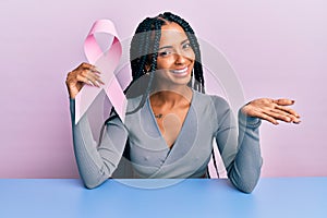 Beautiful hispanic woman holding pink cancer ribbon celebrating achievement with happy smile and winner expression with raised