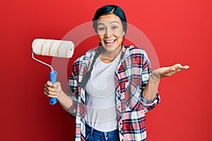 Beautiful hispanic woman holding roller painter celebrating achievement with happy smile and winner expression with raised hand