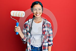 Beautiful hispanic woman holding roller painter looking positive and happy standing and smiling with a confident smile showing