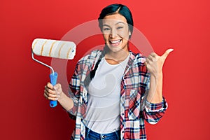 Beautiful hispanic woman holding roller painter pointing thumb up to the side smiling happy with open mouth