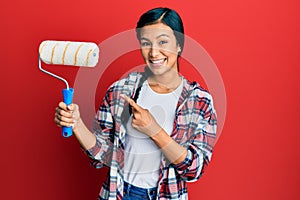 Beautiful hispanic woman holding roller painter smiling happy pointing with hand and finger