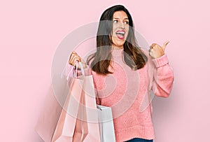 Beautiful hispanic woman holding shopping bags pointing thumb up to the side smiling happy with open mouth