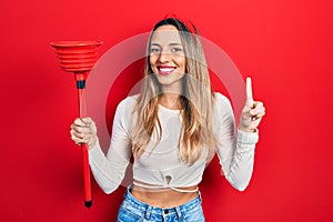 Beautiful hispanic woman holding toilet plunger smiling with an idea or question pointing finger with happy face, number one
