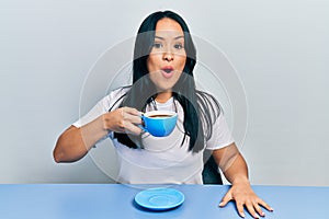 Beautiful hispanic woman with nose piercing drinking a cup coffee scared and amazed with open mouth for surprise, disbelief face