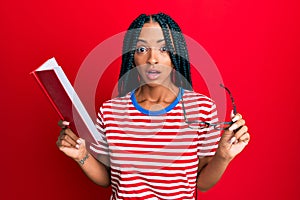 Beautiful hispanic woman reading a book with glasses afraid and shocked with surprise and amazed expression, fear and excited face