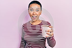 Beautiful hispanic woman with short hair drinking a cup of coffee scared and amazed with open mouth for surprise, disbelief face